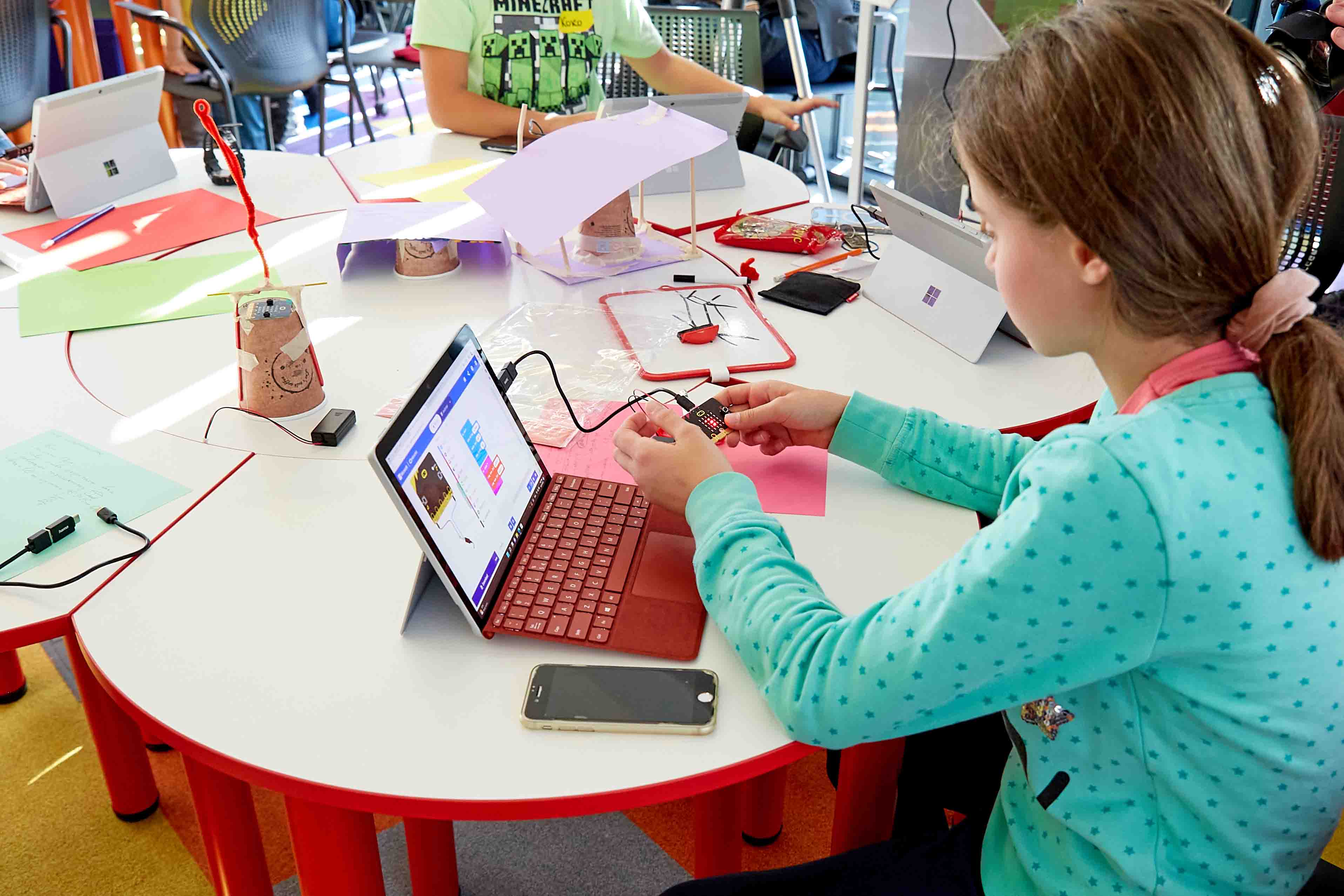 A student sits at a red semi-circular table, working on a laptop and other electronics. Various colored wires, craft materials and a smartphone are spread out on the table in front of her.