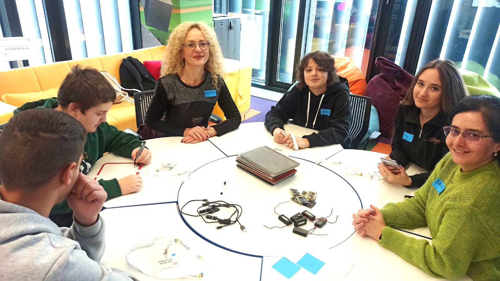 The image shows a group of about 6 students sitting around a circular table with a teacher. They are smiling and appear to be engaged in some kind of learning activity, with various materials on the table in front of them.