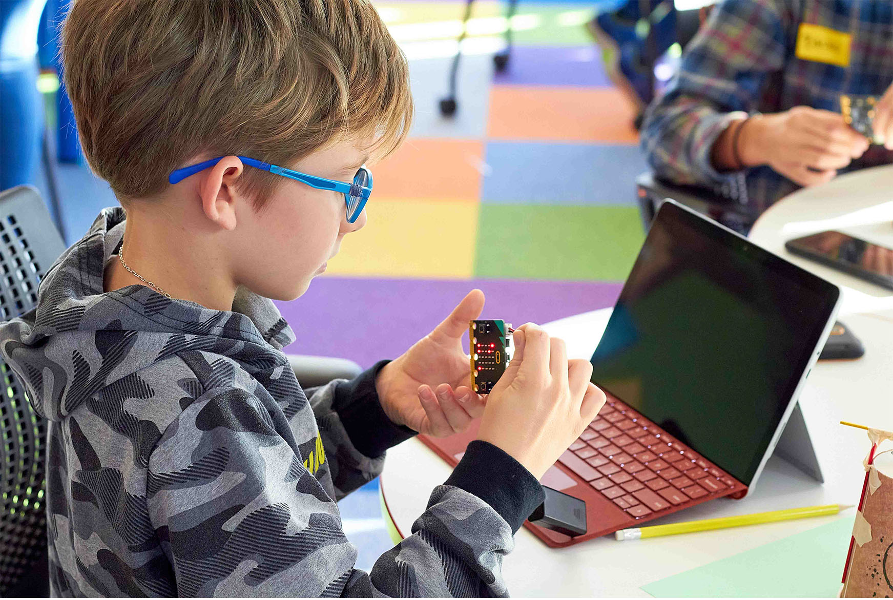 A close-up view of a student, focused on a device while working on a laptop computer in a classroom setting with colorful floor tiles.