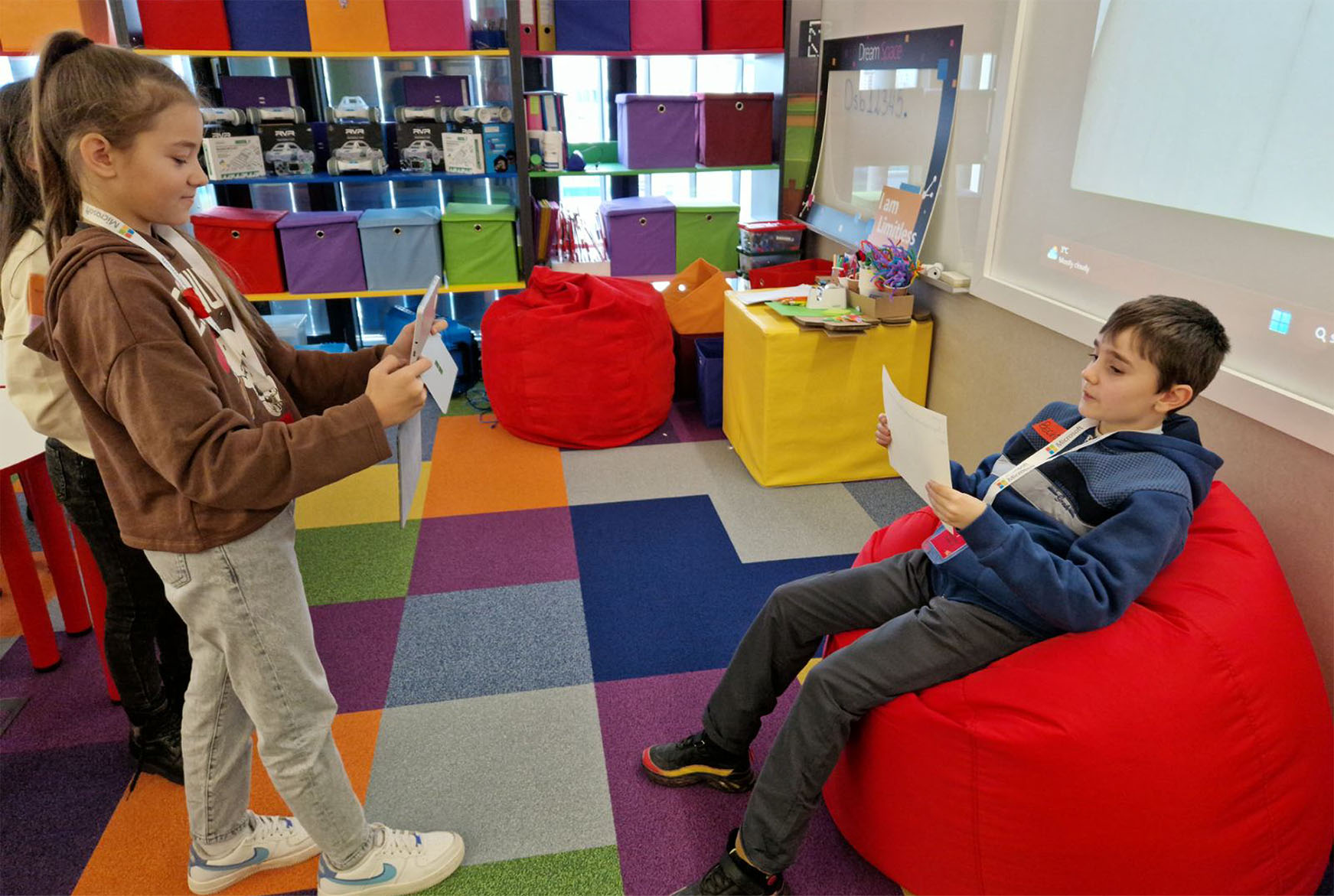 Two students, one standing and holding a tablet, and the other seated on the bean bag with a paper in his hand in a classroom area with colorful cubbies, bins, and a patterned floor.