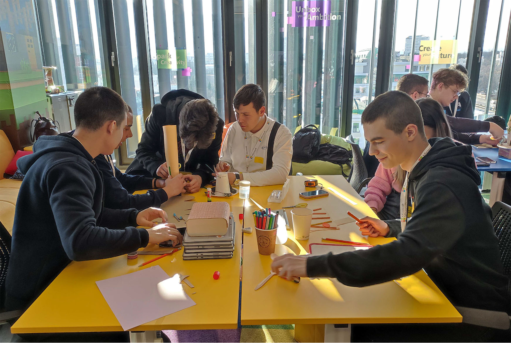 A group of students gathered around a yellow table, engaged in individual work or activities using notebooks, pens, and laptops in a modern, bright learning space with large windows.