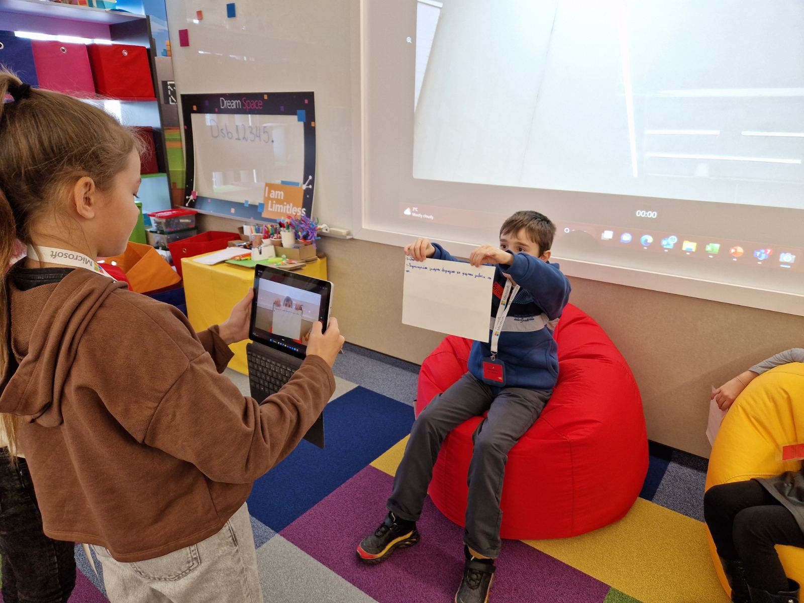 Two young students collaborate while using a tablet and a page. One student is holding a page and other student is scanning it with a tablet. A whiteboard with dream spaces written on it is visible in the background.