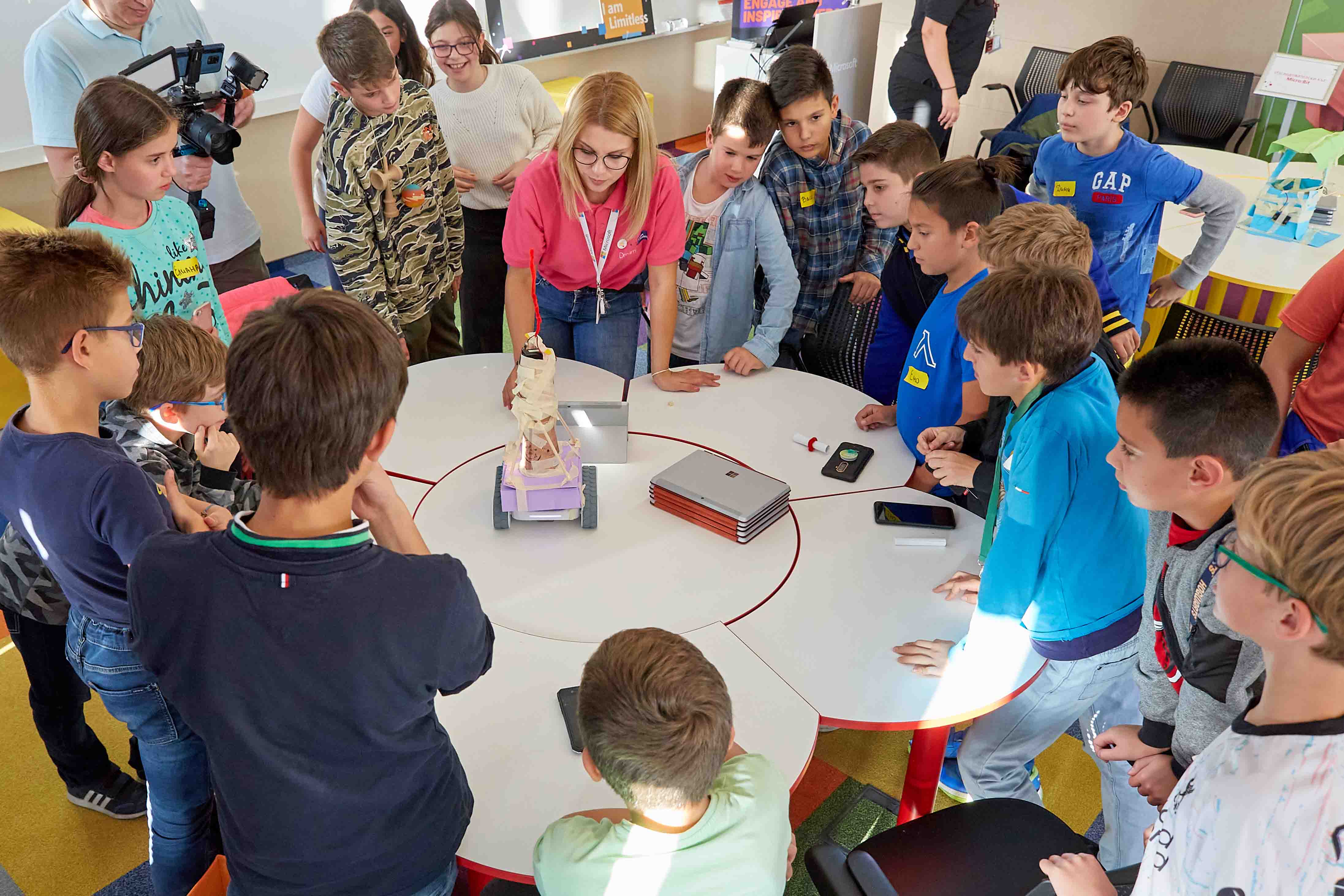 A group of children gather around a white circular table while an educator facilitates an activity. The children are collaborating and working with various electronics and craft supplies on the table.